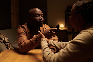 Young irritated man pointing at his wife during quarrel or conflict while sitting by table in front of her and blaming her on his problems