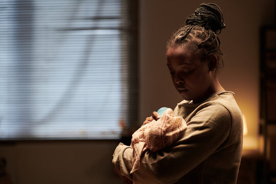 Young African American woman with baby on hands feeding infant with milk while standing in front of camera in home environment