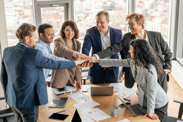 A diverse group of business professionals stand around a conference table, their hands stacked together in a gesture of teamwork and unity. The office is modern and bright