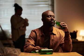 Young African American man with alcohol addiction sitting by table in front of his wife feeding...