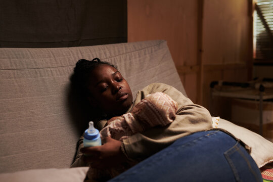 Young sick and tired African American single mother with bottle of milk resting on couch in front of camera and looking at newborn baby