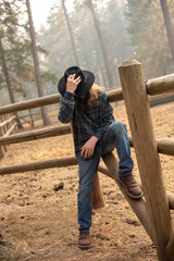 young man with long hair, cowboy hat, flannel shirt, and jeans, sitting on corral fence