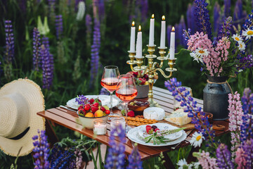 Simple countryside beautiful wedding decor, surprise romantic date outdoors, garden arrangement with lupins flowers on a meadow. Fruits, rose wine, burning candles. Sunset, candlelight, golden hour.