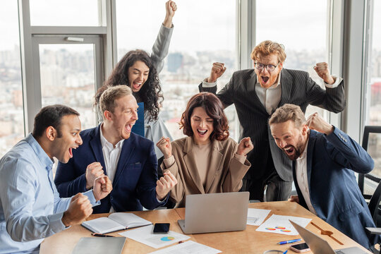 A group of six colleagues in business attire celebrate a success in a modern office with large windows. They are all excited and raise their fists in the air