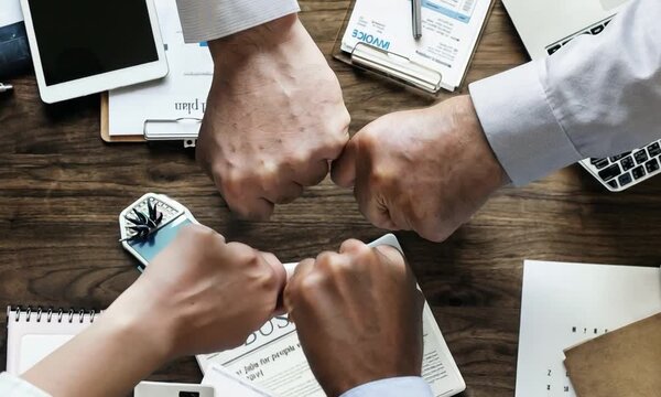A close-up image of four hands coming together in a fist bump over a wooden desk with office supplies. This symbolizes teamwork, collaboration, and unity in a professional setting.
