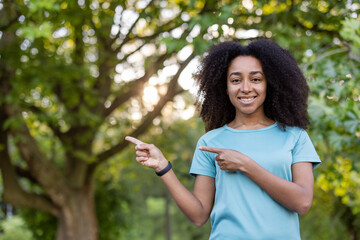 Smiling African American woman in blue shirt pointing sideways in park setting with trees in background. Concept of happiness, outdoors, and positive vibes.