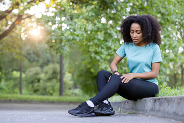 African American woman in sportswear sitting on roadside in park holding knee with a phone in hand. Concept of injury, exercise, and pain during workout.