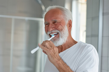 a white-haired, bearded elderly man brushing his teeth in the bathroom, smiling and looking at himself in the mirror