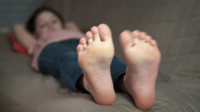 Child feet touch each other. A view of young girl massaging her bare feet on the sofa.