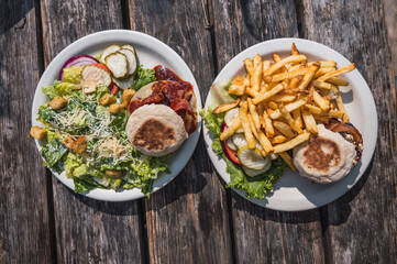 Burger with melted cheese, bacon and beef, accompanied by salad and pickles, and burger with blue cheese, bacon and beef accompanied by French fries.