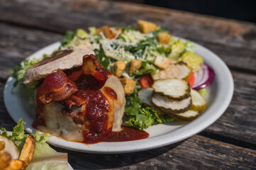 Hamburger with melted cheese, bacon and beef, accompanied by salad and pickles, typical American food, served on a white plate on a rustic wooden table.