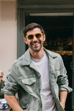 Smiling man wearing yellow sunglasses and a green jacket outdoors