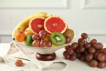 Glass vase with different fresh fruits and grapes on white table, closeup