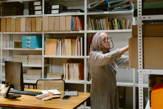 Senior woman in office looking for documents folder on bookshelf