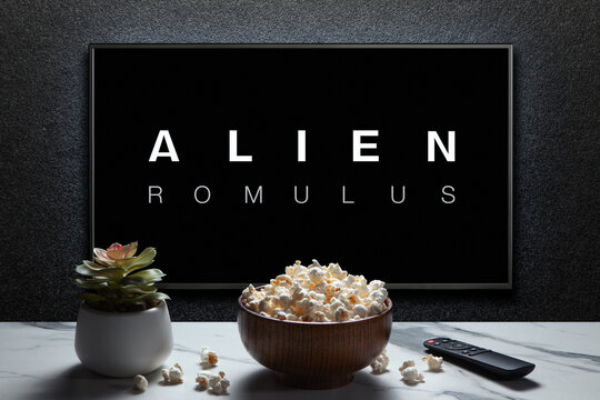 Alien Romulus trailer or movie on TV screen. TV with remote control, popcorn bowl and home plant.