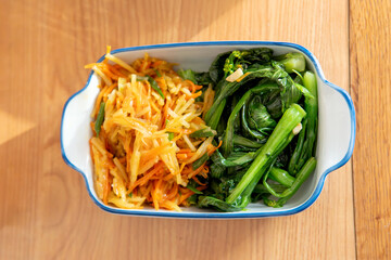A healthy lunch for a Chinese worker in Asia, featuring grilled chicken and pork with potatoes, choy sum, and other vegetables, all illuminated by natural sunlight, making the meal appear very appetiz