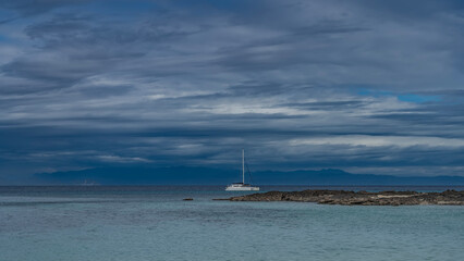 Fototapeta premium A serene seascape. The white yacht is anchored in the calm turquoise ocean. A tall mast against a background of blue sky and clouds. A rocky island. A mountain range in the distance. Madagascar. 