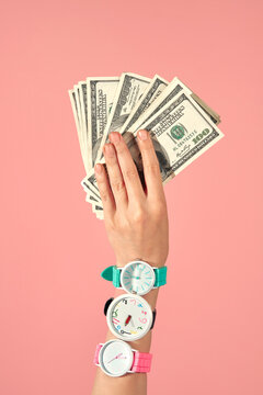 Hand Holding US Dollar Bills With Three Wristwatches on Pink Backdrop.