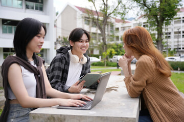 Three students are working together on a project outdoors, using a laptop and a book while sitting at a table