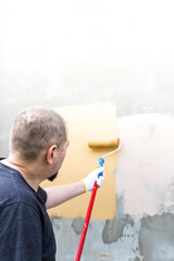 A man is painting a wall using a paint roller attached to a long handle. He is wearing a white...