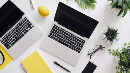 Modern workspace with two laptops, yellow notepad, glasses, smartphone, and plants on a white desk. Top view, minimalist office setup.