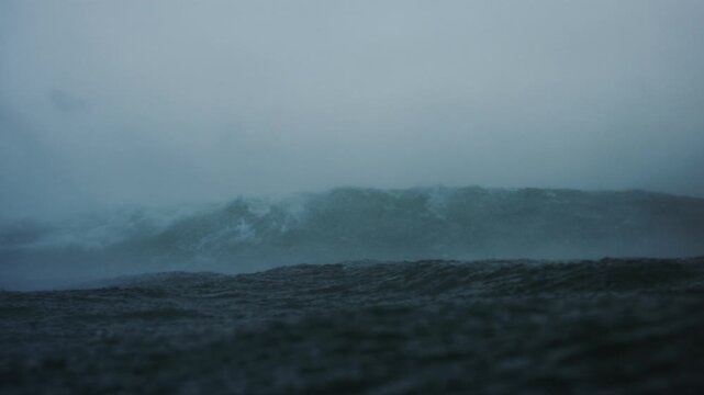 Mist spray rises from top of wave, view from behind in water at surface of ocean on stormy grey day, slow motion