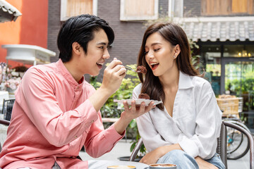 Young asian man feeding happy girlfriend with tasty chocolate cake sitting at table with coffee at date in restaurant