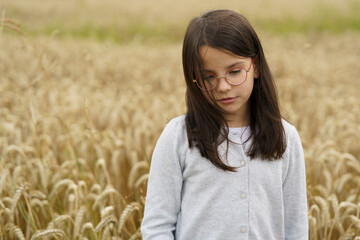 Little cute emotional girl in glasses in the middle of a field with ripe wheat. Concept of young farming and childhood