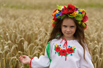 A small sad girl in a colorful wreath and embroidered shirt stands in the middle of a ripe wheat...