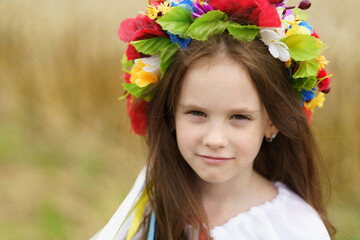 Naklejka premium A cute little emotional girl in a traditional embroidered shirt and with a wreath stands in the middle of a field with ripe wheat. The concept of unity with traditions and nature