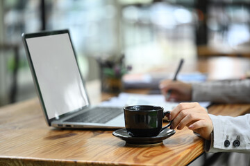 Hands of female worker holding cup of coffee, using laptop at wooden desk in coffee shop
