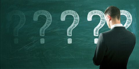 Back view of thoughtful young european male with chalk drawn question marks on blackboard background. Solution, education and knowledge concept.