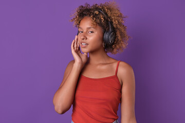 A stylish young woman, wearing headphones, smiles and delicately touches her cheek, immersed in the rhythm of her favorite tunes.