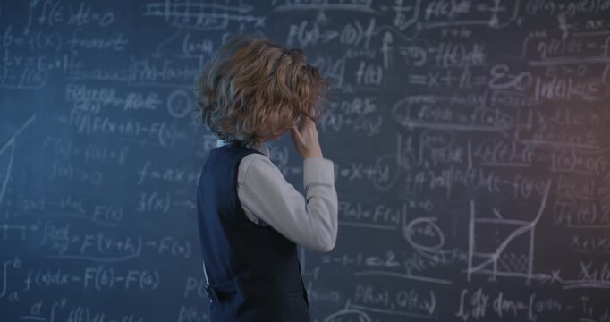 Slow motion of clever little boy standing in classroom and looking at chalkboard with formulas studying algebra alone. Childhood and education concept.