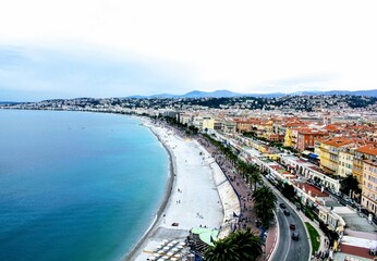 A breathtaking aerial view of a coastal city featuring a curved shoreline with turquoise waters, a bustling promenade, and vibrant rooftops set against a backdrop of distant hills.