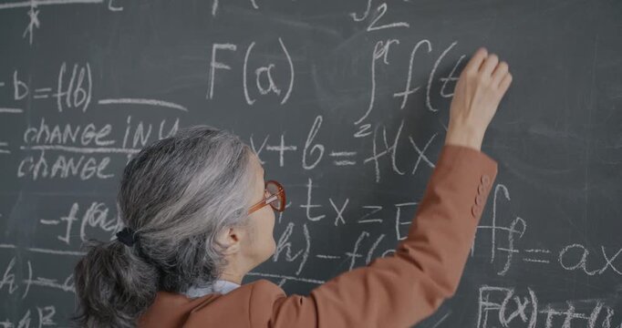 High school teacher senior woman writing formula on chalkboard and speaking teaching algebra in class. Education and people concept.