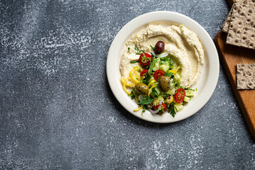 Greek hummus loaded with fresh cucumbers, olives, tomatoes, pepper,  onions and herbs on plate on dark background, space for text, flat lay, close-up