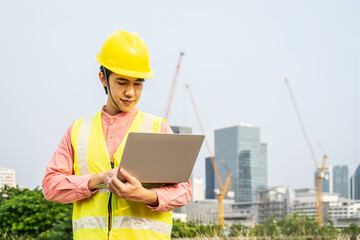 Engineer or construction worker Asian man is holding the laptop and working concentrated at building plan, Hard hat help protect with operation