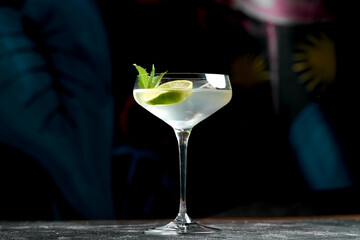 A classic mojito cocktail in a coupe glass with mint leaves and a lime wedge. The drink is served on a dark, moody background.