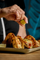 A close-up of a woman's hand squeezing fresh lime juice over three tacos on a ceramic plate.