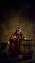 Obraz premium Man, medieval monk in monastic robes sitting in dark cellar, looking inside wooden mug of beer, leaning on barrels with onion and bread appetizers. Concept of comparison of eras, history, brewery
