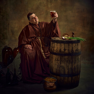 Medieval monk, man lifting pint of ale against dark vintage background. Ancient art of abbey brewing in serene monastic setting. Concept of comparison of eras, history, brewery, traditions