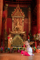 Obraz premium A little girl sits in front of a statue of Buddha. The statue is surrounded by flowers and candles. The girl is wearing a white dress and a pink headband. The scene is peaceful and serene