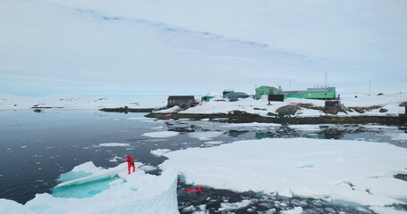 Scientists explore melting glaciers in Antarctica. Two man in orange wetsuits take samples standing on melt ice floe ocean water, climate change problem. Polar station expedition. Aerial drone flight