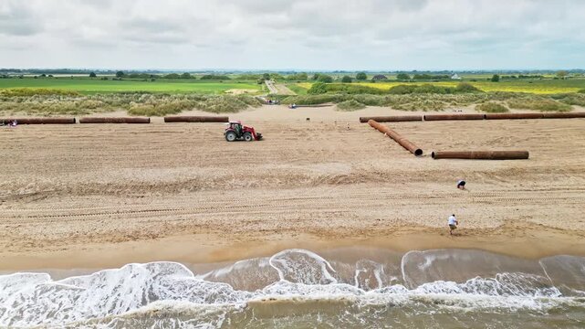 sandy beach with gentle waves, captured by an aerial drone as plant machinery and tractors move irrigation pipework along the coastline in a bustling construction scene.