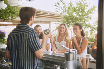 Group of friends relaxing with cocktails at outdoor bar, engaging in lively conversation with male bartender. Outdoor drinks. Concept of friendship, party, leisure, celebration, relaxation