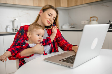 Mother multi-tasking, holding baby infant and using computer laptop at home. Candid authentic and real life mom working and parenting