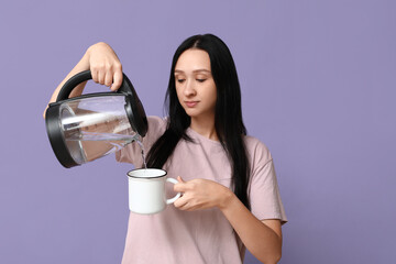 Beautiful young woman pouring hot water from modern electric kettle into mug on lilac background