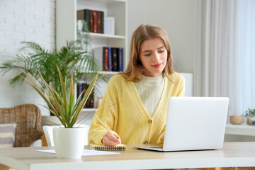 Fototapeta premium Young woman working remotely on laptop at home office desk with notebook and pen, she is writing in a notebook with a pen, demonstrating focus and productivity