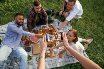 Taking photo by using smartphone. Group of friends are having picnic on the field with food in eco...